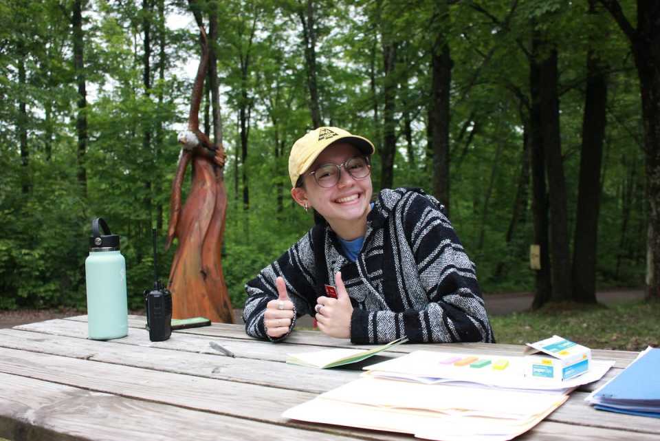 A girl leans over a wooden park table and smiles, with the forest visible behind